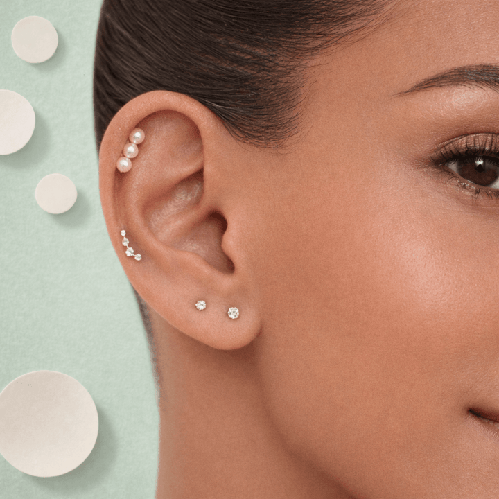 Close-up of a woman's ear with multiple earrings against a light green background with circular patterns.